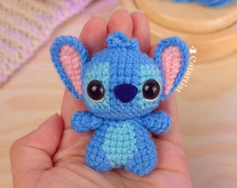 May include: A handmade blue crochet Stitch doll with large black eyes and pink inner ears. The small plush toy is held in a hand, showcasing its detailed craftsmanship. A red crochet heart is visible in the background.
