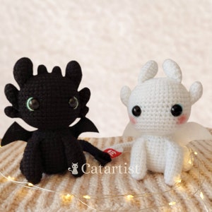 May include: Two crocheted plush toys, one black and one white, resembling dragons from the movie How to Train Your Dragon. The black dragon has a green eye and the white dragon has a blue eye.