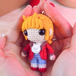 Peut inclure: Une poupée au crochet faite à la main avec des cheveux orange, de grands yeux noirs et une veste rouge. La poupée porte un short bleu et des chaussures rouges. La poupée est tenue dans des mains ouvertes. Le texte "Catartist" est visible.