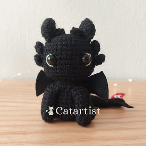 Puede incluir: Juguete de peluche de dragón negro de crochet con ojos verdes y un ala de fieltro negro. El dragón está sentado sobre una superficie de madera. El texto "Catartist" es visible en el juguete.
