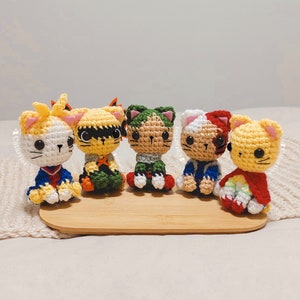 Puede incluir: Cinco peluches de gato tejidos a crochet, cada uno con un disfraz diferente inspirado en personajes populares de anime. Los peluches están sentados sobre una superficie de madera.