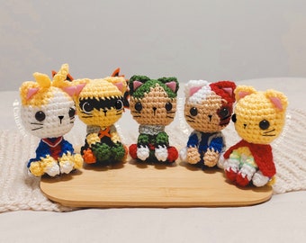 May include: Five crocheted cat plushies, each wearing a different costume inspired by popular anime characters. The plushies are sitting on a wooden surface.