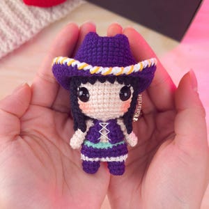 Peut inclure: Poupée au crochet faite à la main avec un chapeau de cow-boy violet, des cheveux noirs et une robe violette. La poupée a de grands yeux noirs et est tenue dans des mains ouvertes. Le chapeau a une bordure blanche et jaune. La robe de la poupée a une rayure vert clair et des accents blancs.