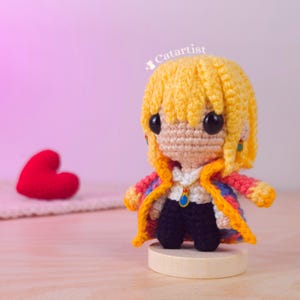 Puede incluir: Muñeca de ganchillo hecha a mano con cabello amarillo, que lleva un abrigo colorido y pantalones negros. La muñeca tiene ojos negros grandes y un pequeño colgante. Un objeto en forma de corazón rojo está en el fondo. La muñeca está sobre una base de madera.