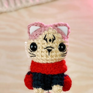 May include: A handmade crochet cat figurine with a pink and cream-colored head, black eyes, and a red scarf. The cat wears a navy blue outfit with red accents. The figurine is attached to a pink stick.