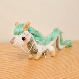 Puede incluir: Un juguete de dragón de crochet con un cuerpo blanco y gris, pelo verde claro y cuernos marrones. El dragón está sentado sobre una superficie de madera.