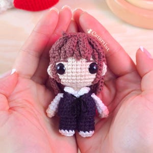 Könnte beinhalten: Eine handgefertigte Häkelpuppe mit braunen Haaren und großen schwarzen Augen. Die Puppe trägt ein dunkelblaues Outfit mit weißen Akzenten. Die Puppe wird in offenen Händen gehalten. Der Name des Künstlers, Catartist, ist sichtbar.