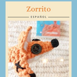 May include: A crocheted fox toy with black eyes and a black button nose. The fox is orange and white and has black accents on its paws and ears. The fox is lying on a white fuzzy surface.