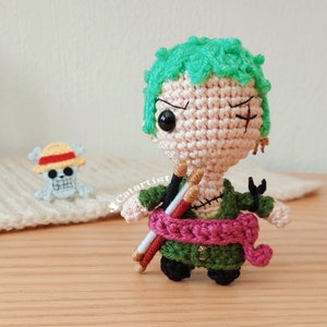 Puede incluir: Una figura amigurumi de crochet de un personaje de pelo verde con una faja rosa y dos palos. El personaje tiene un parche en el ojo negro y una cicatriz cosida. La figura está de pie sobre una superficie de madera.