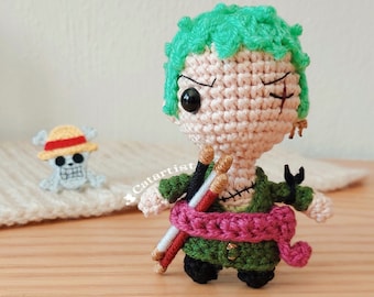 May include: A crocheted amigurumi figure of a green-haired character with a pink sash and two sticks. The character has a black eye patch and a stitched-on scar. The figure is standing on a wooden surface.