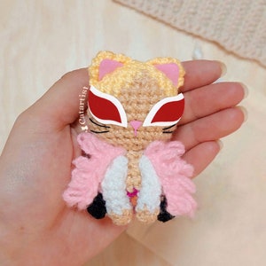 Puede incluir: Un gato de crochet con una chaqueta rosa y blanca y gafas de sol rojas y blancas. El gato sostiene una flor rosa en su pata.