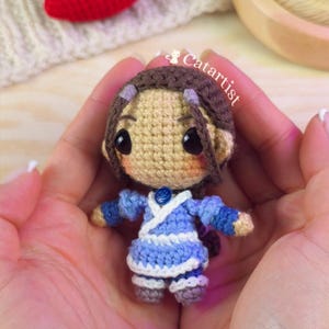 May include: A small crochet doll with brown hair, large black eyes, and pink cheeks. The doll wears a light blue and white outfit with a small blue circular emblem at the neck. The text Catartist is visible near the top of the doll's head.