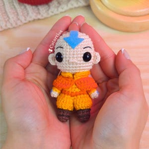 Pode incluir: Uma pequena boneca de crochê de um personagem careca com uma seta azul na testa, vestindo uma roupa laranja e amarela com botas marrons. A boneca tem olhos pretos grandes e o texto Catartist está impresso em branco na lateral da cabeça.