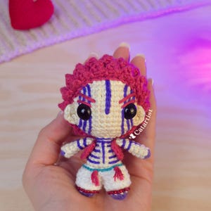 Puede incluir: Muñeca de ganchillo hecha a mano con una cara color crema y pantalones blancos. La muñeca tiene pelo y detalles rosas, con detalles azules y morados en la cara y la ropa. La muñeca se sostiene en una mano. Un corazón rojo está en el fondo.