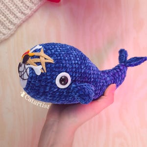 Puede incluir: Un peluche de ballena azul tejido a ganchillo con un diseño de temática pirata. La ballena presenta una calavera y huesos cruzados, un parche en el ojo y el texto "Catartist". El peluche se sostiene en una mano.