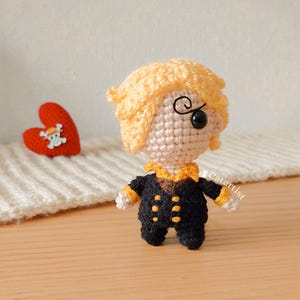 Puede incluir: Una muñeca de crochet con cabello amarillo, un traje negro con botones dorados y un solo ojo negro. La muñeca está de pie sobre una superficie de madera con un corazón rojo con una calavera y huesos cruzados en el fondo.