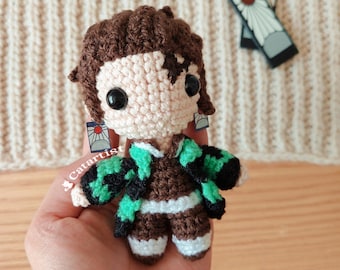 May include: Crocheted anime character with brown hair, green and black patterned jacket, and white pants. The character has black eyes and is wearing earrings with a red and white sun design.