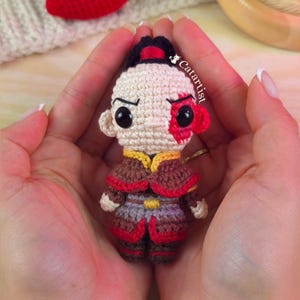 May include: A small crocheted doll featuring a character with a black ponytail, a beige face, and a prominent red scar over the left eye. The figure wears a brown and red outfit with a gold belt detail. The text Catartist is visible on the side.