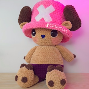 May include: A handmade plush toy of a character wearing a pink hat with a white cross and brown ears. The plush has a tan body, purple shorts, and brown feet. The text "Catartist" is on the hat.