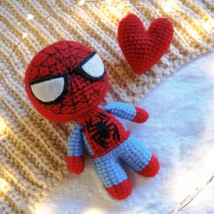 May include: A red and blue crocheted Spiderman toy with a black spider design on the chest, lying on a white fuzzy surface with a red crocheted heart beside it.