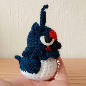 May include: A crocheted blue and white bird with a red and black eye and a long, thin, blue tail. The bird is sitting on a hand.