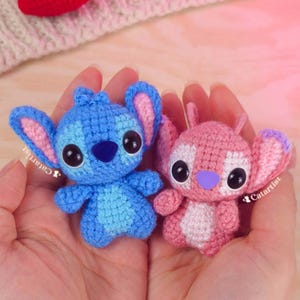 Puede incluir: Dos pequeñas figuras de ganchillo de una criatura azul y rosa con aspecto de extraterrestre, con grandes ojos negros y grandes orejas. La figura azul tiene detalles azules y rosas, mientras que la figura rosa tiene detalles rosas y blancos. Las figuras están en manos de una persona.