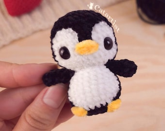 May include: A handmade crochet penguin with a black head, wings, and feet, and a white body. The penguin has large black eyes and a yellow beak. The text "Catartist" is visible on the penguin. A small red heart is in the background.