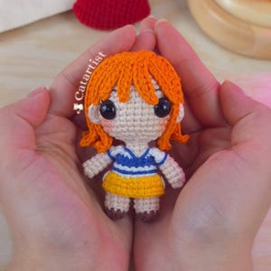 Puede incluir: Muñeca de ganchillo hecha a mano con pelo naranja, ojos negros grandes y un atuendo azul y amarillo. La muñeca se sostiene en manos abiertas, con un corazón rojo de ganchillo en el fondo. La muñeca mide unos 8 cm de alto.