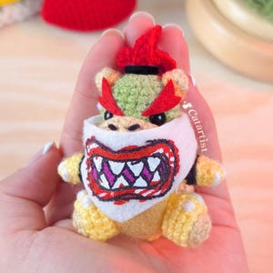 May include: A handmade crochet Bowser figure, featuring a green head, red horns and hair, and a white bib with a detailed mouth design. The figure has yellow and tan accents, and black eyes. The figure is held in a hand.