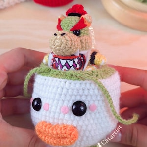 Op de afbeelding: Gehaakte Bowser Jr. amigurumi pop. De pop heeft een witte body met een groene rand, zwarte ogen, roze wangen en een oranje snavel. Bowser Jr. zit erbovenop, met een rode schaal en kroon. Handgemaakt.
