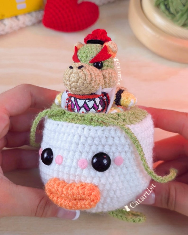 Op de afbeelding: Gehaakte Bowser Jr. amigurumi pop. De pop heeft een witte body met een groene rand, zwarte ogen, roze wangen en een oranje snavel. Bowser Jr. zit erbovenop, met een rode schaal en kroon. Handgemaakt.