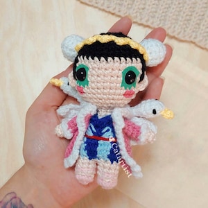 May include: A crocheted doll with pink and white clothing, black hair, and green eyes. The doll is holding a white crocheted bird with a yellow beak.