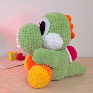 Puede incluir: Un peluche de Yoshi verde hecho a mano con ganchillo, con detalles blancos, naranjas y rojos. El peluche tiene ojos negros grandes y una barriga blanca. El texto "Catartist" es visible.