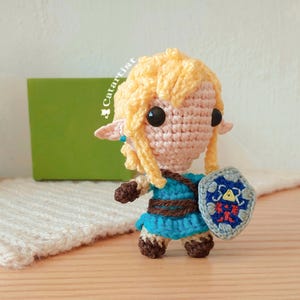 Puede incluir: Un juguete de peluche tejido a crochet de un personaje con cabello rubio, ropa azul y un escudo con un diseño triangular. El juguete está de pie sobre una superficie de madera.