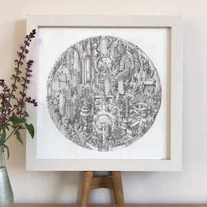 May include: A black and white drawing of a city skyline, rendered in a circular shape. The drawing is framed in a white frame and is displayed on a wooden easel.