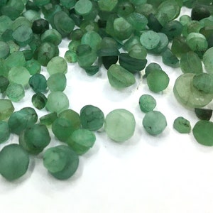 May include: A large collection of small, loose emerald gemstones in various shades of green. The stones are faceted with irregular, rounded shapes and varying levels of translucency, scattered across a plain white surface in a dense pile.