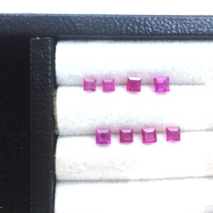 以下が含まれることがあります： 2列に並べられた8個の小さな正方形カットの鮮やかなピンク色の宝石。石はファセットカットされており、明るく半透明な色をしています。白いパッド入りのディスプレイ表面に配置されています。