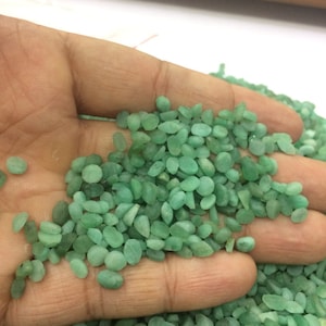 Puede incluir: Primer plano de una mano que sostiene una colección de pequeñas piedras verdes redondeadas. Las piedras son lisas y pulidas, con un aspecto ligeramente translúcido. Las piedras están dispersas sobre una mano, con más piedras visibles en el fondo. Las piedras tienen un tamaño y forma uniformes.