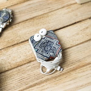 Puede incluir: Una pequeña bolsa de tela azul y blanca con estampado con auriculares blancos en su interior. La bolsa está sobre una superficie de madera con un reloj y una cámara.
