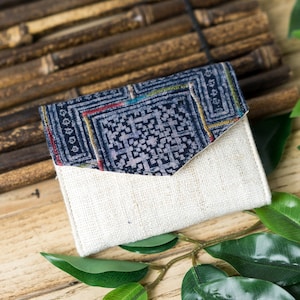 May include: A small, white linen wallet with a blue and gold patterned flap. The wallet is sitting on a woven bamboo mat.