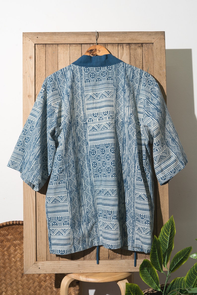 May include: A blue and white patterned kimono with a tie closure. The kimono is hanging on a wooden hanger against a wooden background.