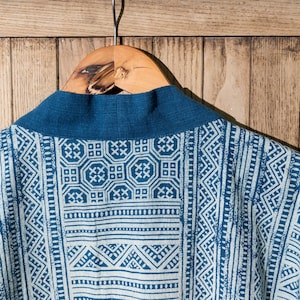 May include: A blue and white patterned kimono with a dark blue collar. The kimono is hanging on a wooden hanger against a woodgrain background.