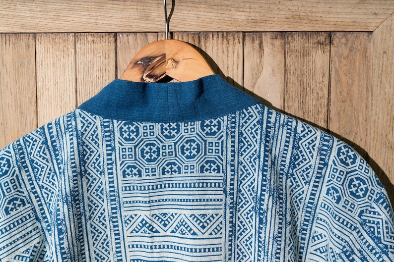 May include: A blue and white patterned kimono with a dark blue collar. The kimono is hanging on a wooden hanger against a woodgrain background.