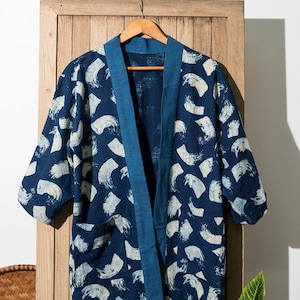 May include: A blue and white patterned kimono-style jacket with a blue trim. The jacket is hanging on a wooden hanger against a white wall.