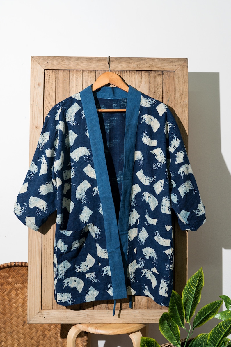 May include: A blue and white patterned kimono-style jacket with a blue trim. The jacket is hanging on a wooden hanger against a white wall.