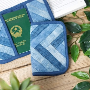 Puede incluir: Dos porta pasaportes de mezclilla azul con un pasaporte verde dentro de uno de ellos. Los porta pasaportes están hechos de mezclilla de retazos y tienen un borde azul. Los porta pasaportes están sobre una superficie de madera con hojas verdes.