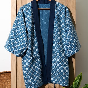 May include: A blue and white patterned kimono with a dark blue trim. The kimono is hanging on a wooden hanger against a wooden background.