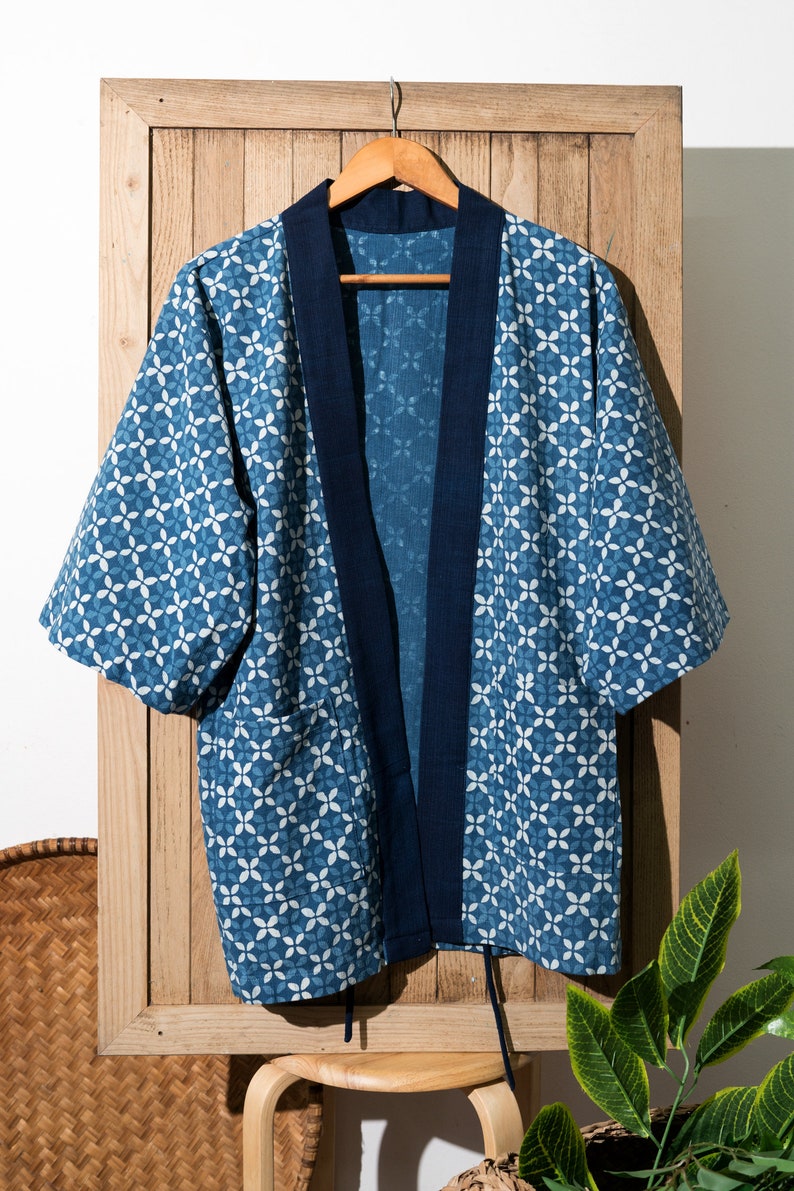 May include: A blue and white patterned kimono with a dark blue trim. The kimono is hanging on a wooden hanger against a wooden background.