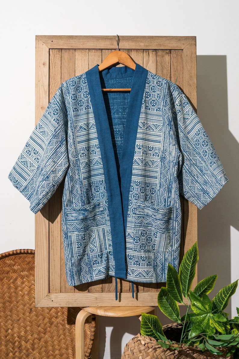 May include: A blue and white patterned kimono with a blue trim and a tie closure. The kimono is hanging on a wooden hanger against a white wall.