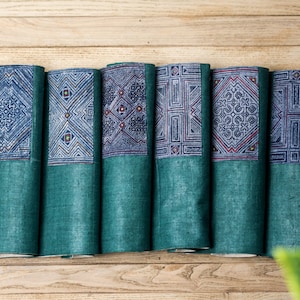 May include: Six teal blue fabric placemats with a blue and white geometric pattern. The placemats are folded and arranged in a row on a wooden surface.